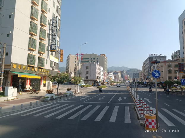 Eine Stadtstraße mit Gebäuden, Fahrzeugen, Straßenlaternen, Schildern, Absperrpollern, Bäumen, Pflanzen, Namensschildern, Strommasten, Kabeln, Hügeln und Himmel im Hintergrund.