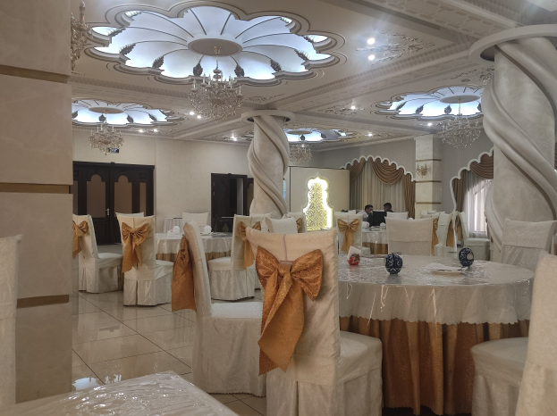 Ein großer Festsaal mit Tischen und Stühlen, die mit Tüchern drapiert und mit verschiedenen Gegenständen geschmückt sind, mit Wänden, die mit Türen und Vorhängen dekoriert sind, und einer Decke, die mit Lichtern und Kronleuchtern geschmückt ist.