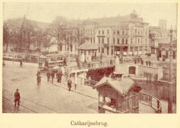 Ein altes Foto einer Stadtstraße mit Menschen, die umgeben sind von Gebäuden, Bäumen, Pfählen und einer Brücke, mit Text unten.