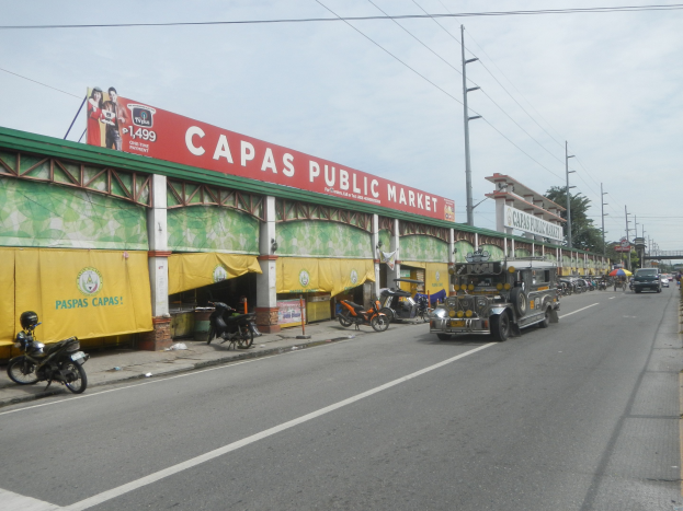 Eine belebte Straße mit Fahrzeugen, einem Gehweg, Strommästen, Gebäuden, Bäumen und einem bewölkten Himmel, mit einem Gebäude mit der Aufschrift "Capas Public Market" im Vordergrund.