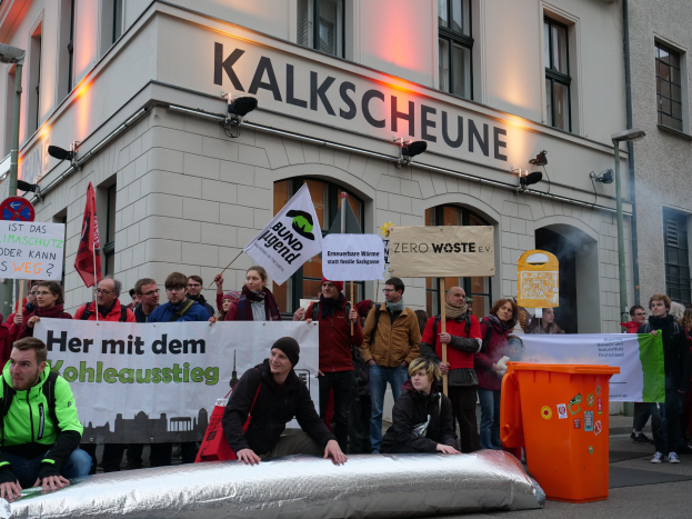 Eine Gruppe von Menschen mit Schildern und Transparenten steht vor einem Gebäude, mit zwei Personen im Vordergrund und einem Müllcontainer rechts, während einer Protestaktion in Deutschland.