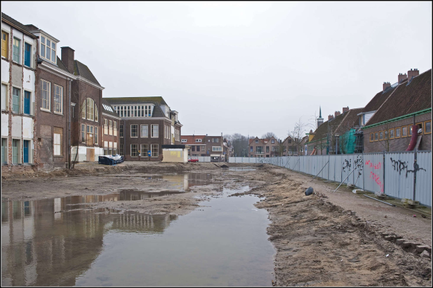 Flutende Stadtstraße mit Wasser auf dem Boden, ein Zaun auf der rechten Seite, Gebäude mit Fenstern auf der linken Seite, Bäume im Hintergrund und Himmel darüber.