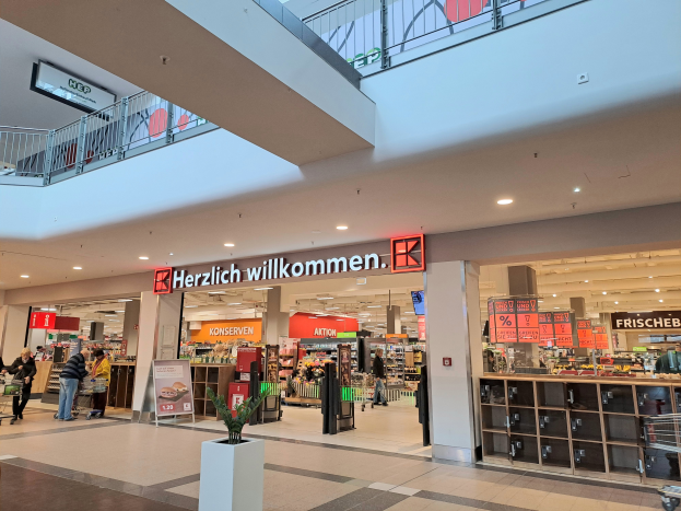 Das Innere eines Einkaufszentrums mit Menschen, die herumlaufen, Geschäften, Schildern mit Text, Lampen, Geländern und anderen Gegenständen, mit einem Topf mit einer Pflanze unten und dem Text "Herzlich willkommen" auf den Schildern.