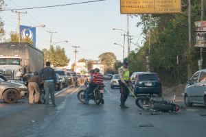 Eine Gruppe von Menschen umringt ein verunglücktes Motorrad am Straßenrand mit mehreren Fahrzeugen, darunter ein Lastwagen, und einer Hintergrundlandschaft aus Bäumen, Pfosten, Lampen und Schildern unter dem Himmel.
