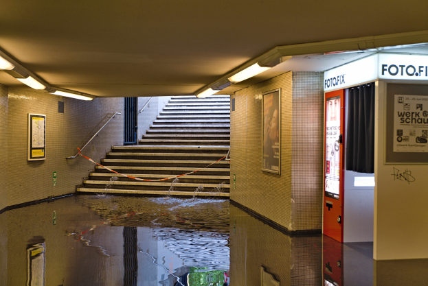 Eine überflutete U-Bahn-Station in Berlin, Deutschland, mit Wasser auf dem Boden, Treppen mit Geländern, Foto-Rahmen an den Wänden, Lampen an der Decke und einem Automaten.