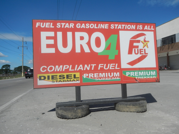 Eine Tankstelle mit einem "Euro4 Fuel"-Schild im Vordergrund, Fahrzeuge auf der Straße, Strommäste, Bäume, ein Gebäude auf der rechten Seite und ein sichtbarer Himmel.