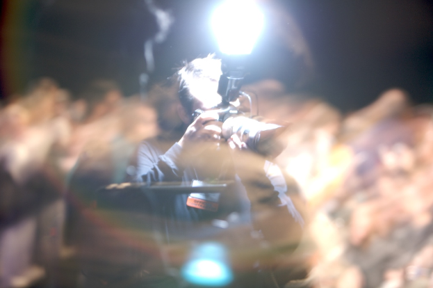 Eine Person mit einer Digitalkamera in der Hand, vor einem unscharfen Hintergrund und einem Blitz oben auf dem Bild.