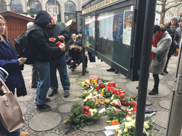 Eine Gruppe von Menschen versammelt sich an einer Bushaltestelle, die mit Blumen und Kerzen geschmückt ist, mit einem Gebäude mit Bögen und Fenstern, Bäumen und einem Schuppen im Hintergrund, um an ein tragisches Schussereignis in Berlin, Deutschland, zu erinnern.
