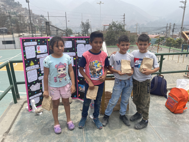 Gruppe von Kindern auf einem Basketballfeld mit Papiertüten in den Händen, Tüten und Gegenstände rechts und eine Tafel mit Text links, vor einem Hintergrund aus Zaun, Pfosten, Laternen, Bäumen, Gebäuden, Hügeln und Himmel.