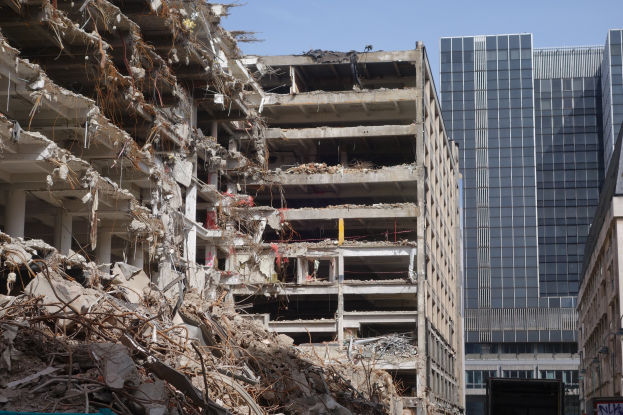 Ein großes Gebäude wird in einer Stadt abgerissen, umgeben von intakten Gebäuden, Straßeninfrastruktur, Fahrzeugen, Bäumen und einem klaren Himmel.
