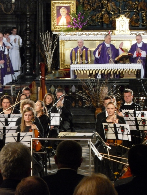 Eine Gruppe von Menschen, die Geigen spielen, mit Noten vor sich, mit anderen Individuen im Vordergrund, drei Kirchenväter dahinter mit einer Bibel und ein Photo-Rahmen mit Verzierungen im Hintergrund.