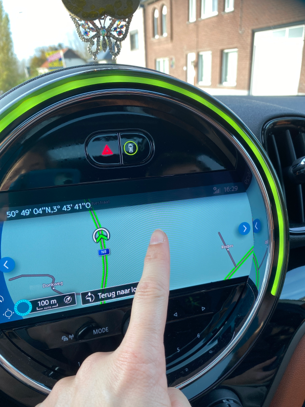 Eine Person zeigt auf ein GPS-Gerät auf dem Armaturenbrett eines Autos, mit Gebäuden, Bäumen und einem klaren blauen Himmel im Hintergrund.