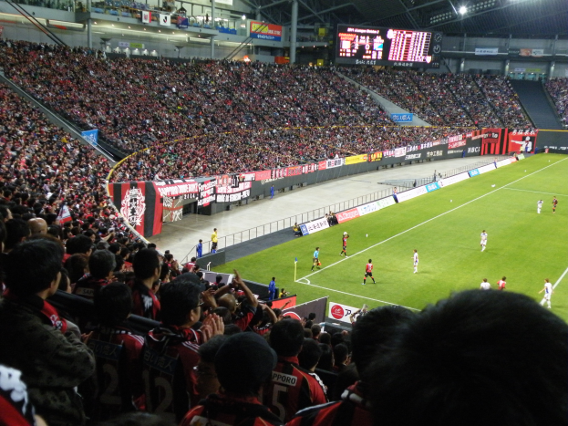 Ein Fußballspiel in einem großen Stadion mit Spielern auf dem Feld, Zuschauern in den Rängen, Bannern, einem Zaun, einem Anzeigebildschirm, Metallpfosten und einem Dach mit Deckenleuchten.