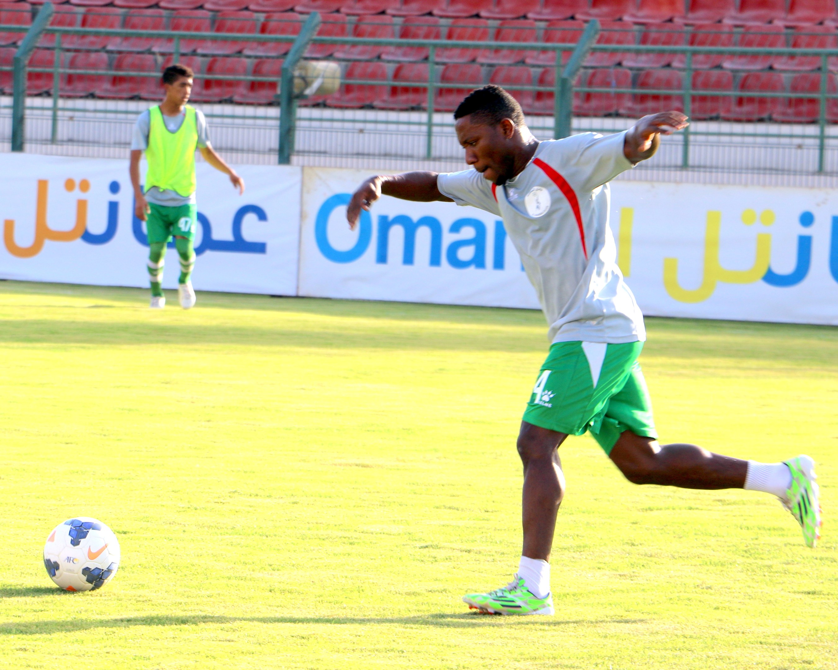 Ein Mann kickt einen Fußball auf einem Rasenfeld in einem Stadion, mit zwei Personen in der Nähe und Bannern, Geländern und Stühlen im Hintergrund.