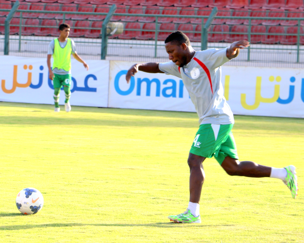 Ein Mann kickt einen Fußball auf einem Rasenfeld in einem Stadion, mit zwei Personen in der Nähe und Bannern, Geländern und Stühlen im Hintergrund.