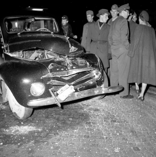 Eine Schwarz-Weiss-Szene eines beschädigten Autos am Strassenrand, mit einer Gruppe von Menschen drumherum, die besorgt aussehen.