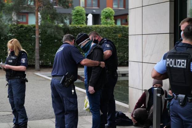 Deutsche Polizeibeamte stehen in einer Gruppe vor einem Gebäude, während eine Frau ein Buch und einen Stift hält und andere sitzen oder stehen in der Nähe, umgeben von Grünflächen und städtischen Strukturen.