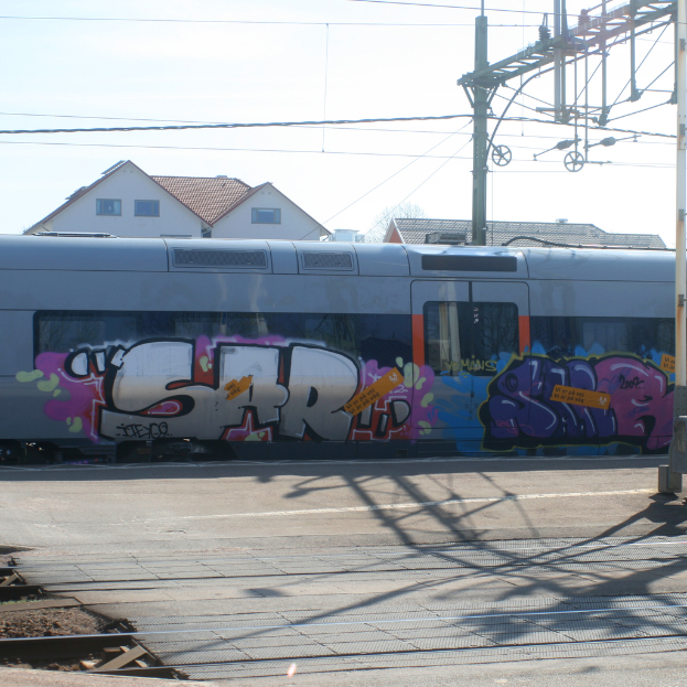 Ein mit Graffiti bedeckter Zug mit sichtbaren Kabeln und Häusern mit Fenstern im Hintergrund.