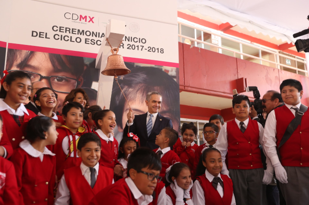 Eine Gruppe von Kindern in roten Uniformen steht vor einem Banner mit Text und einem Bild einer Person, mit einer Glocke in der Mitte und einem Fotografen auf der rechten Seite, vor einem Schulgebäude in Mexiko-Stadt während eines CDMX-Events.