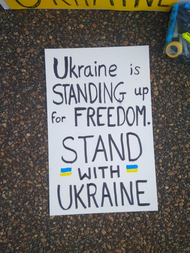 Weißer Zettel mit "Ukraine steht für Freiheit, steht mit Ukraine" Text auf dem Boden vor einer gelben Fahne, mit einer Plastikabdeckung mit verschiedenen Gegenständen rechts daneben.