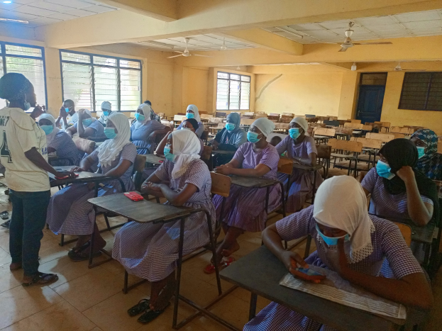 Ein Klassenraum in Nigeria mit Schülern, die Gesichtsmasken tragen und auf Bänken und dem Boden sitzen, mit Büchern und Gegenständen auf Tischen, Fenstern, einer Tür, einer Wand, einer Decke mit Lampen und Ventilatoren im Hintergrund.