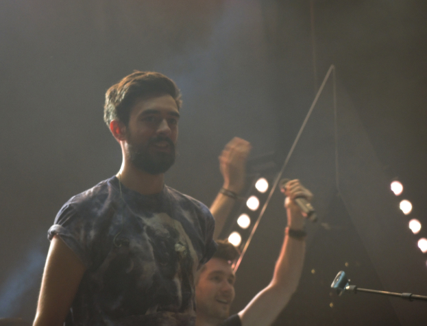 Ein Mann in einem T-Shirt steht und lacht, mit einem anderen lachenden Mann, Lichtern, Rauch, einem Mikrofon und anderem Equipment im Hintergrund.