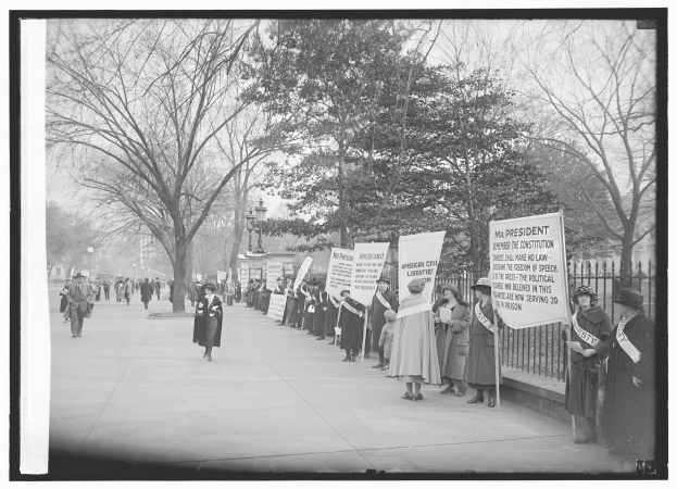 Eine Gruppe von Suffragetten marschiert auf einem baumgesäumten Bürgersteig, hält Schilder, mit einem Zaun auf der rechten Seite, dargestellt in Schwarz-Weiß.