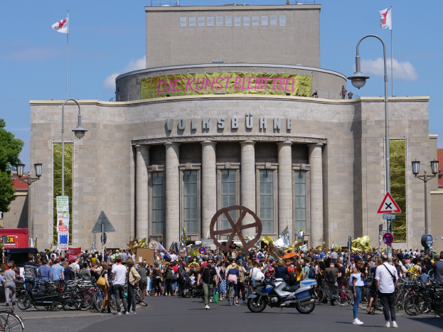 Eine große Gruppe von Menschen steht vor einem Gebäude mit Säulen und Text, umgeben von Straßenmasten, Straßenlaternen, Schildern, Motorradfahrern, Kraftfahrzeugen, Bäumen und einem bewölktem Himmel bei einer Demonstration in Berlin, Deutschland.