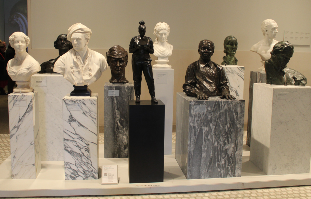 Eine Gruppe von detailreichen Büsten-Skulpturen, die in einem Museum ausgestellt sind, mit ein paar Menschen im Hintergrund und einer Wand dahinter.