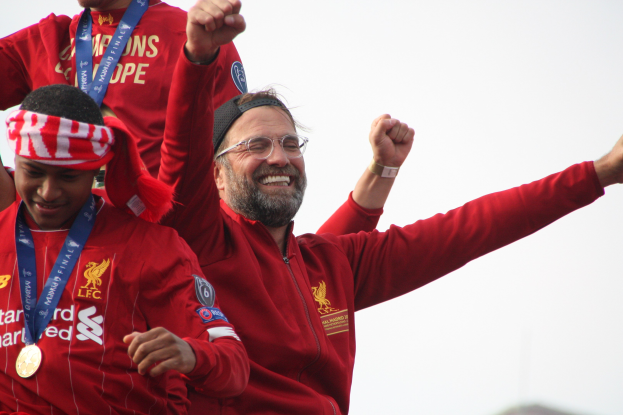 Liverpool-Trainer Jurgen Klopp feiert mit Spielern und Staff nach dem Gewinn der UEFA Champions League, trägt eine rote Jacke mit zwei medaillentragenden Individuen neben ihm gegen einen Himmel-Hintergrund.