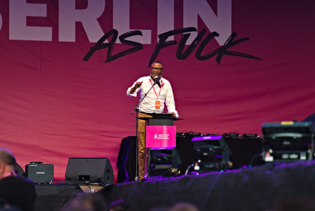 Ein Mann in einem weißen Hemd und braunen Hosen steht auf einer Bühne und hält ein Mikrofon, während er eine Menge anspricht. Im Hintergrund ist ein pinkes Banner mit der Aufschrift 'Berlin as Fuck' zu sehen.