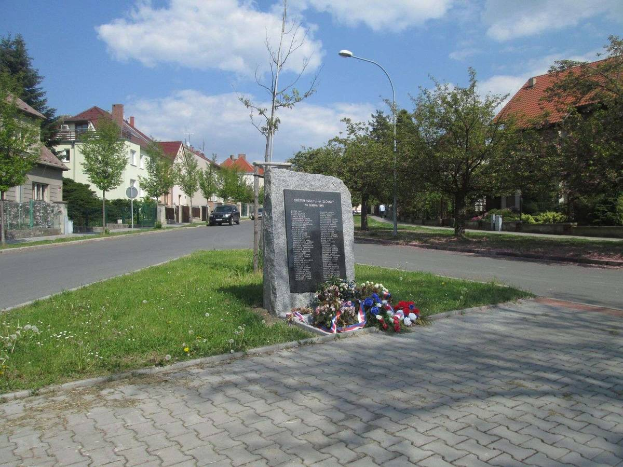 Ein Gedenkstein in einer Stra{\u}en, geschm"{u}ckt mit Blumen, umgeben von Gr"{u}n, Laternen und Fahrzeugen, mit Geb"{a}uden und einem bew"{o}lktem Himmel im Hintergrund, gewidmet den Holocaust-Opfern.