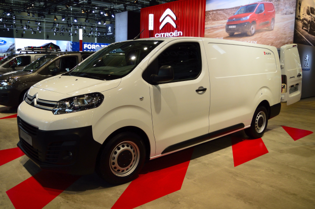 Ein weißer Citroën Berlingo Van wird auf einer Autauschau präsentiert, umgeben von schwarzer Schlafzimmer-Möbeln, während die Wände mit Brettern und Lichtern geschmückt sind.