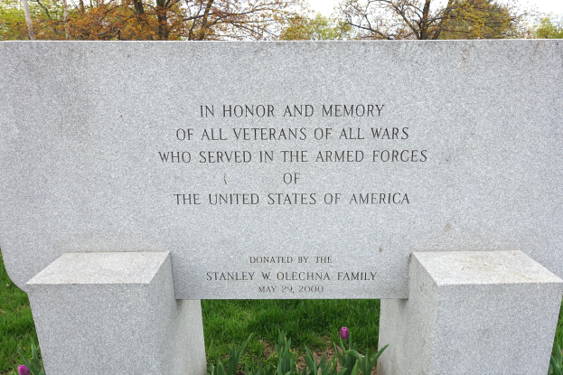 Ein Gedenkstein mit einer Inschrift, die alle US-Veteranen ehrt, umgeben von Pflanzen, Blumen, Gras, Bäumen und unter einem bewölkten Himmel.