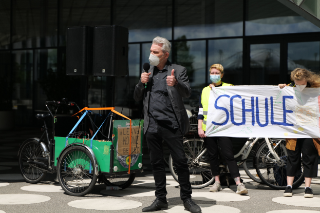 Ein Mann mit Maske hält ein Mikrofon vor einer Gruppe von Menschen, einige halten ein Banner mit Text, Fahrräder, ein Fahrzeug mit einer Kiste und Lautsprecher, vor einem Gebäude mit Glaswänden im Hintergrund.