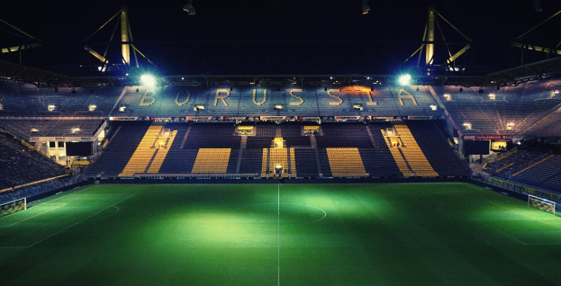Ein Fußballfeld bei Nacht, erhellt von hellen Stadionlichtern, mit einigen Zuschauern auf den Tribünen und saftig grünem Rasen auf dem Feld.