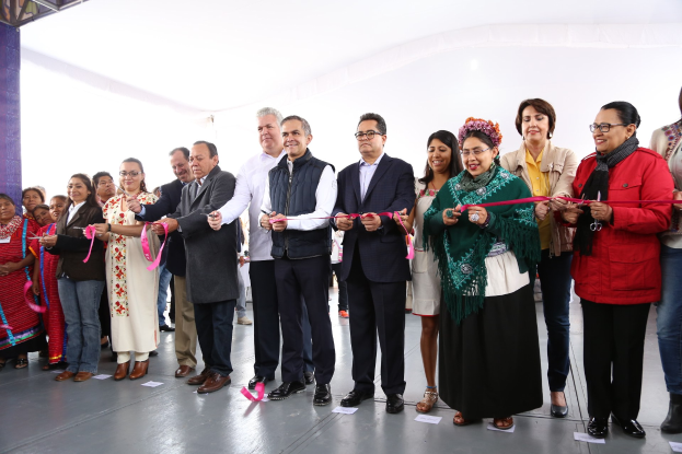 Eine Gruppe von Menschen bei einer Scherenschnitte-Zeremonie, die vor einer Menge mit einer Säule links daneben, verstreuten Papieren auf dem Boden und einem weißen Tuch im Hintergrund stehen.