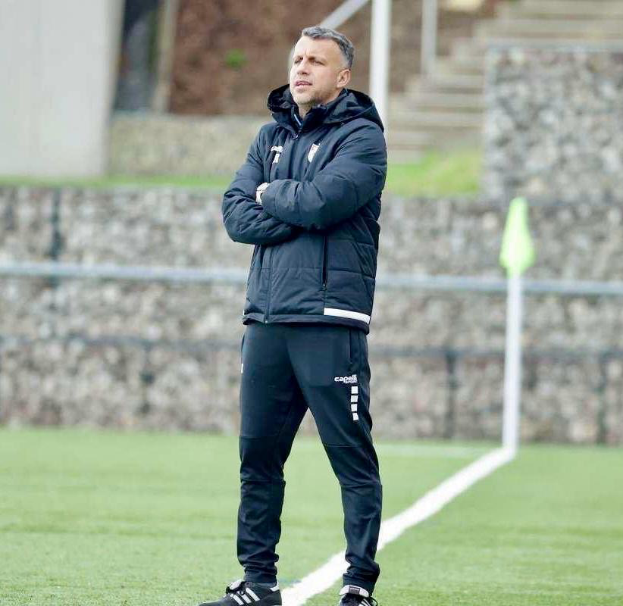 Ein Mann steht auf einem Fußballfeld mit verschränkten Armen und trägt eine schwarze Jacke und Hose, im Hintergrund gibt es einen Zaun, Treppen und eine Wand.