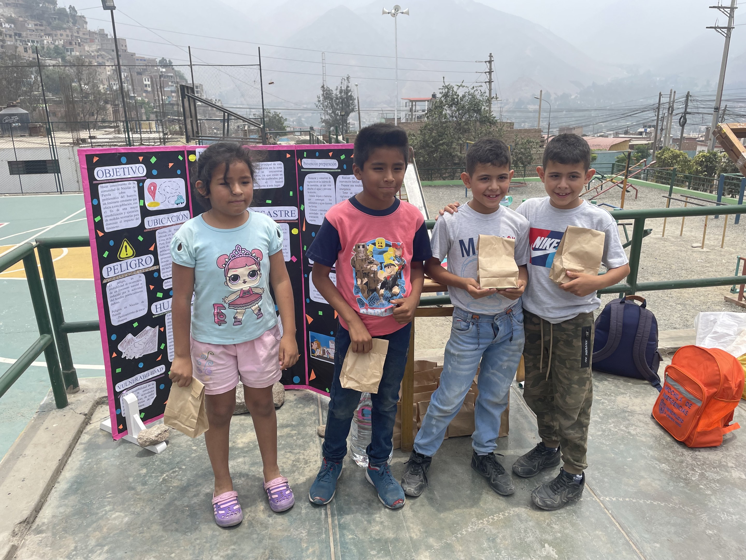 Eine Gruppe von Kindern steht auf einem Basketballfeld und hält Papiertüten in den Händen, mit Tüten und Gegenständen rechts daneben und einer Tafel mit Text links daneben, vor einem Hintergrund aus Zaun, Pfosten, Laternen, Bäumen, Gebäuden, Hügeln und Himmel.