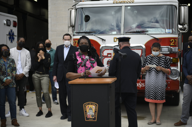 Bürgermeisterin von Chicago Lori Lightfoot steht an einem Rednerpult mit Mikrofonen und Papieren, spricht auf einer Pressekonferenz vor einem Löschfahrzeug, umgeben von maskierten Personen, mit einer beleuchteten Wand im Hintergrund.