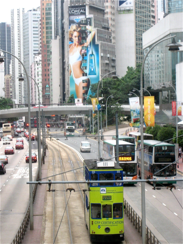 Eine belebte Stadtstraße mit Doppeldeckerbussen, Autos und anderen Fahrzeugen, umgeben von hohen Gebäuden, Bäumen, Pfählen, Laternen, Schildern, Bannern und einer Brücke unter einem klaren blauen Himmel, mit Text unten.