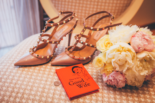 Ein Tisch zeigt ein Buch mit dem Titel "Wie werde ich eine gute Ehefrau", einen Blumenstrauß und ein Paar Valentino-Schuhe, vor einem Vorhanghintergrund.