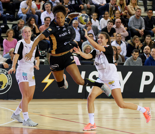 Eine Gruppe von Frauen spielt Handball auf einem Feld mit einem Ball in der Mitte, Zuschauern im Hintergrund und einer Fahne mit Text und Logos.