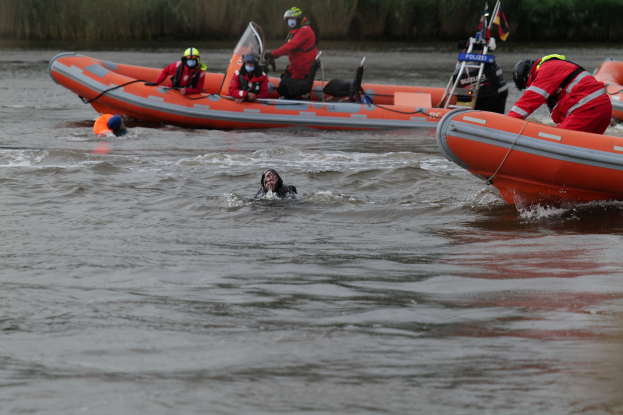 Eine Gruppe von Menschen in einem aufblasbaren Boot auf einem Fluss, mit zwei Personen im Wasser im Vordergrund und Vegetation im Hintergrund, alle mit Schwimmwesten und Helmen während einer Rettungsoperation.