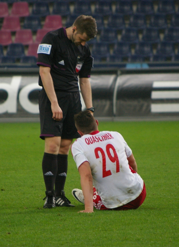 Ein Fussballspieler sitzt neben einem Schiedsrichter auf dem Boden, beide in Sportkleidung, mit Stadiontafeln und Stühlen im Hintergrund.