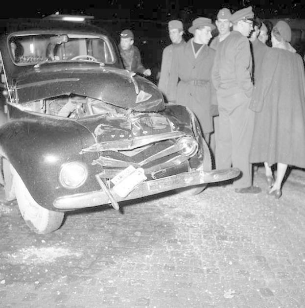 Schwarz-weißes Bild eines schwerbeschädigten Autos am Straßenrand mit eingedellter Motorhaube und zersplitterten Fenstern, umgeben von einer Gruppe von Menschen, die die Szene begutachten.