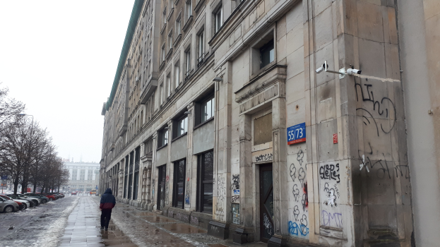 Eine Person, die auf einem Gehweg an einem Gebäude mit Graffiti vorbeigeht, mit parkenden Fahrzeugen, Bäumen und Gebäuden im Hintergrund unter dem Himmel.