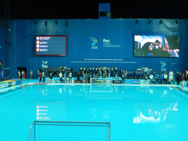 Eine Gruppe von Menschen um ein Hallenbad mit einem Netz am Boden herumstehend, mit einer Wand im Hintergrund, die "FINA World Championships 2015 Kazan"-Text, Logos, Träger und Lichter zeigt.