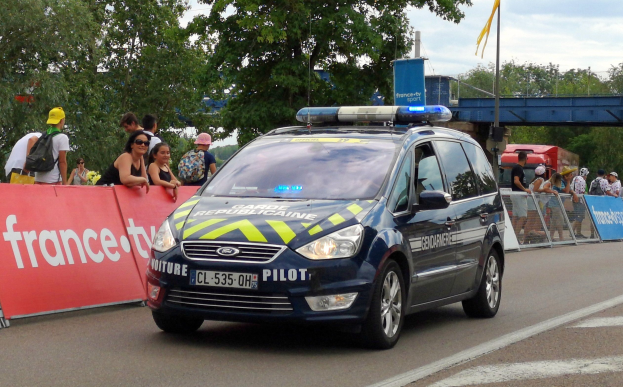 Ein Polizeiwagen fährt auf einer Straße neben einer Menschenmenge, einige tragen Kappen und tägliche Taschen, mit einem Banner links, Geländern mit Bannern dahinter und einem Hintergrund aus Bäumen, einer Brücke, einer Fahne auf einem Mast und einem bewölktem Himmel.
