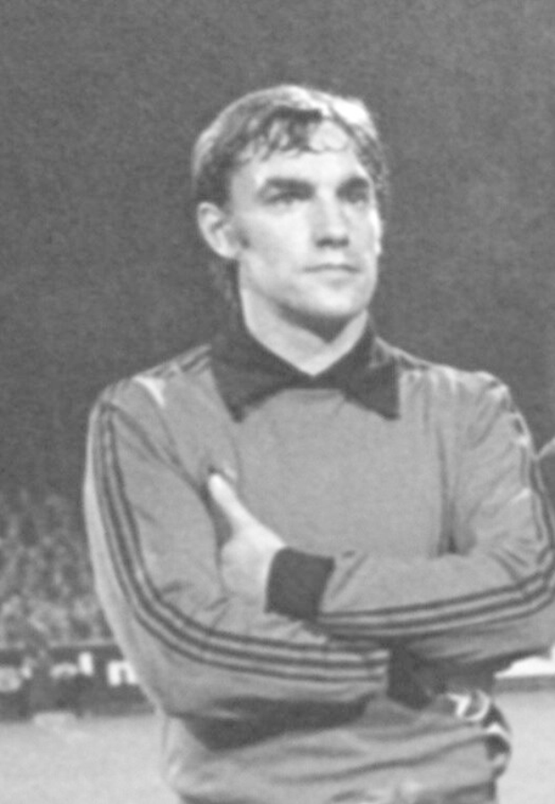Ein Schwarz-Weiß-Foto eines älteren Mannes, der mit verschränkten Armen auf einem Fußballfeld steht und von einer Gruppe Menschen im Hintergrund umgeben ist.